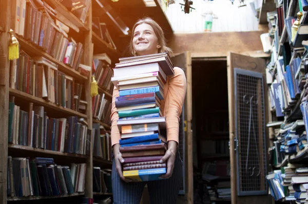 Fille étudiante détient une grande pile de livres et porte beaucoup de littérature dans la bibliothèque, elle se prépare pour l'étude, le vendeur de livres a pris beaucoup de livres dans le contexte d'une librairie — Photo