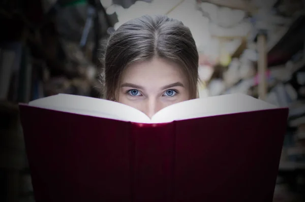 girl student reads a big red book in the library and looks at the camera, she has blue eyes, close-up