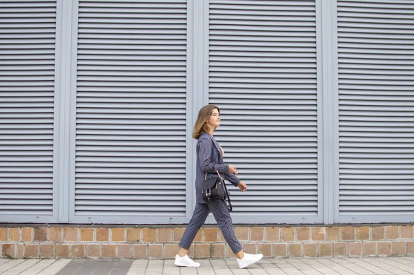 Деловая женщина утром идет на работу в город на фоне стены, девушка в полосатом костюме гуляет по улице — стоковое фото