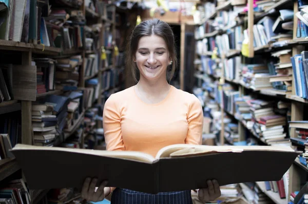 Fille étudiante lit un grand livre dans la bibliothèque, une femme aime lire beaucoup, elle est à la recherche de littérature sur les étagères, la connaissance est le pouvoir, concept — Photo