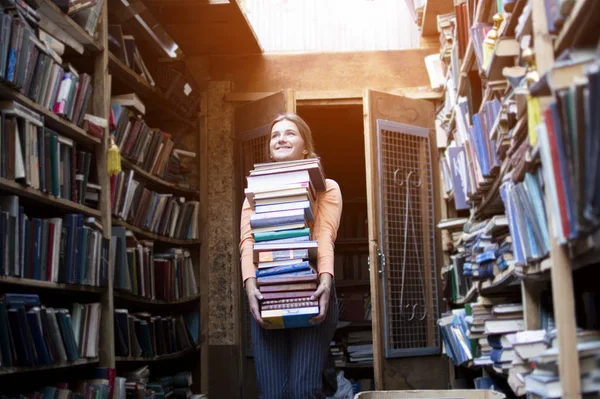 Девушка студентка держит большую пачку книг и несет много литературы в библиотеке, она готовится к учебе, продавец книг взял много книг на фоне книжного магазина — стоковое фото