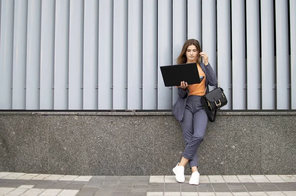 ラップトップを持つビジネスウーマンは、壁の背景に対して市内に滞在し、屋外のコンピュータを使用してストライプのスーツを着た女の子 — ストック写真