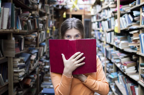 Fille étudiante lit un grand livre rouge dans la bibliothèque et regarde la caméra, elle a les yeux bleus, gros plan — Photo