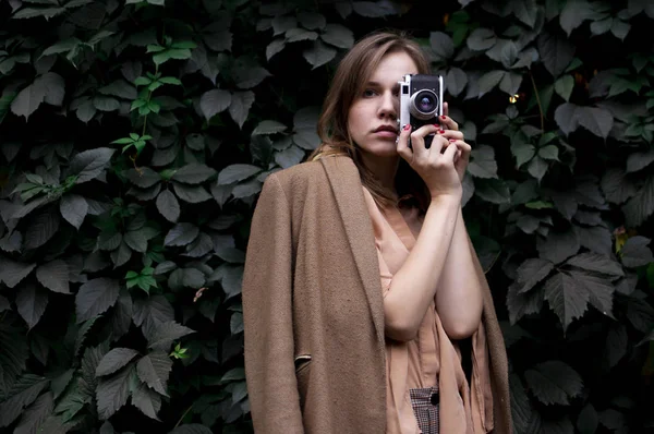 Jovem fotógrafo fica com uma câmera de filme perto de uma parede de folhas na floresta, uma mulher fotografa na natureza — Fotografia de Stock