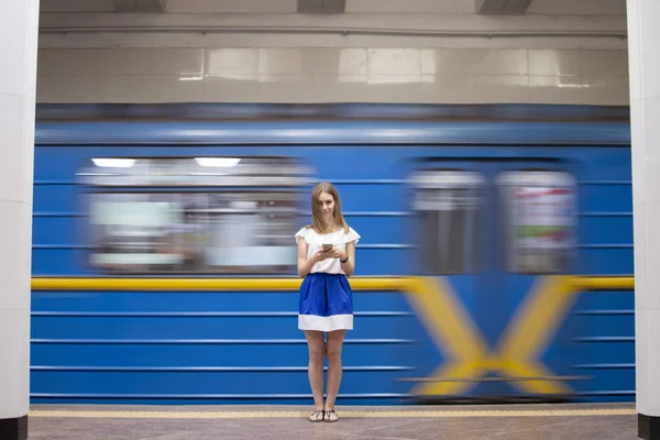 Dziewczyna stoi na platformie metra i słucha muzyki na tle przejeżdżającego pociągu, uczeń korzysta z telefonu w metrze, Kopiuj przestrzeń — Zdjęcie stockowe