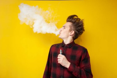 Genç hippi adam sarı izole edilmiş arka planda sigara içiyor, bir duman bulutu salıyor ve elinde elektronik bir sigara tutuyor.