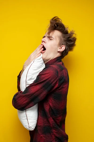 young sleepy tired guy yawns and holds a pillow on yellow isolated background, bad morning, insomnia concept, vertical frame