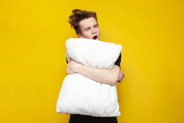 young sleepy tired guy yawns and holds a pillow on yellow isolated background, bad morning, insomnia concept