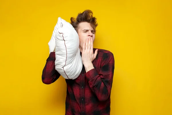 young sleepy tired guy yawns and holds a pillow on yellow isolated background, bad morning, insomnia concept