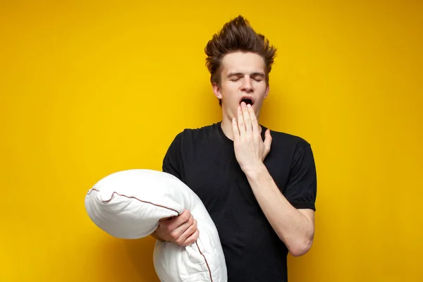 young sleepy tired guy yawns and holds a pillow on yellow isolated background, bad morning, insomnia concept
