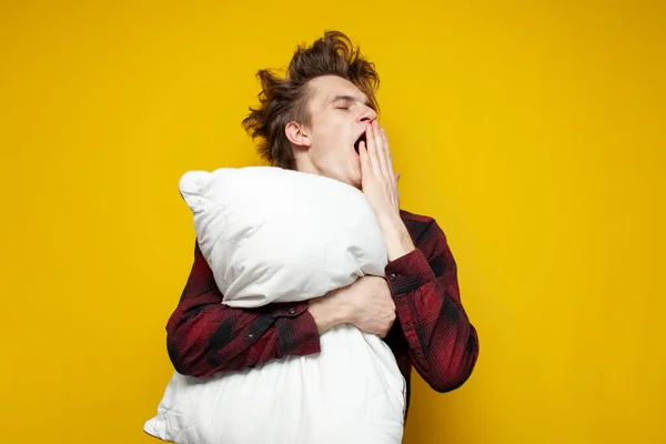 young sleepy tired guy yawns and holds a pillow on yellow isolated background, bad morning, insomnia concept