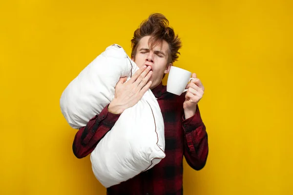 young sleepy tired guy yawns and holds pillow and coffee in the morning on yellow isolated background, bad morning, lack of sleep