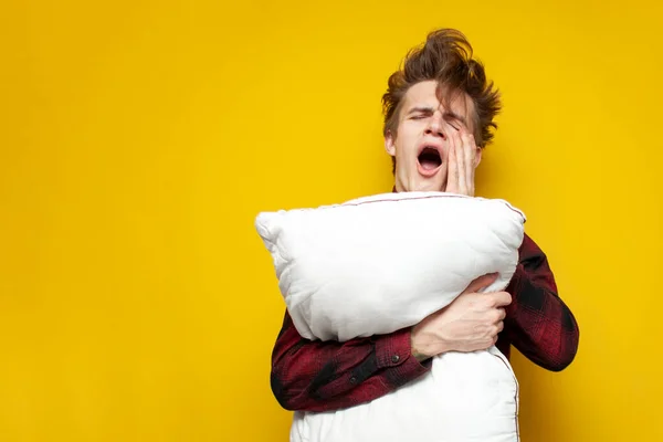 young sleepy tired guy yawns and holds a pillow on yellow isolated background, bad morning, insomnia concept