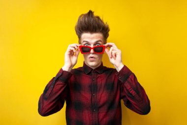 Tuhaf saç stili olan, sarı izole edilmiş arka planda gözlük takan genç, şaşkın bir adam, hipster şaşırır.