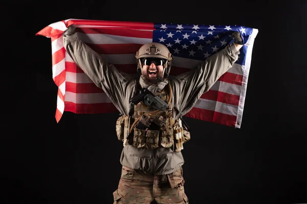 American commando in military equipment holds USA flag at night, modern ranger screams in the dark, elite troops, war concept