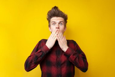 Genç şokta olan adam, elleri sarı izole bir arka planda ağzını kapatıyor, korkmuş adam şaşırıyor.