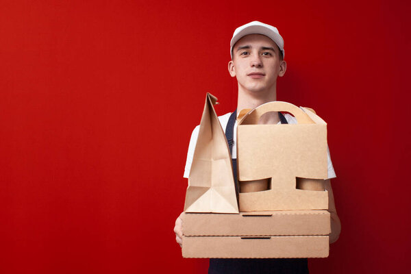 young positive food delivery man on red background, courier guy holds a lot of packaging and smiles, copy space