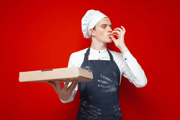 年轻的面包师拿着比萨饼 在红色孤立的背景上表现出很好的品味 他是个厨师 还带着一盒比萨饼 — 图库照片