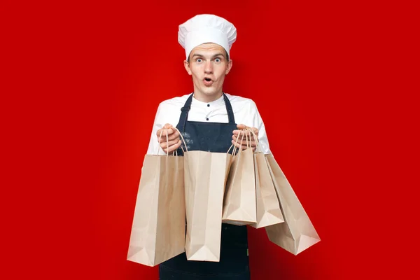 Jovem Cozinheiro Detém Sacos Supermercado Fundo Vermelho Cara Uniforme Chef — Fotografia de Stock