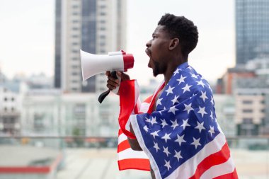 Amerikan bayrağı taşıyan Afro-Amerikan bir adam megafonla bağırıyor ve şehrin arka planına karşı protestolar düzenliyor. Siyahların hayatı kavram meselesidir.