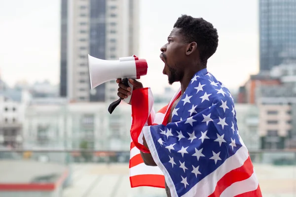 Amerikan bayrağı taşıyan Afro-Amerikan bir adam megafonla bağırıyor ve şehrin arka planına karşı protestolar düzenliyor. Siyahların hayatı kavram meselesidir.