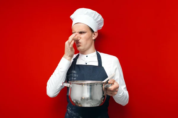 身着制服的年轻厨师端着一道难吃的菜 对着一个红色孤立的背景 臭味难闻的概念闭口 — 图库照片