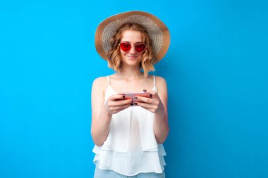 Güneş şapkalı ve gözlüklü genç bir kız mavi bir arka planda akıllı telefon kullanıyor, yaz kıyafetleri içindeki bir kadın telefona bakıyor.