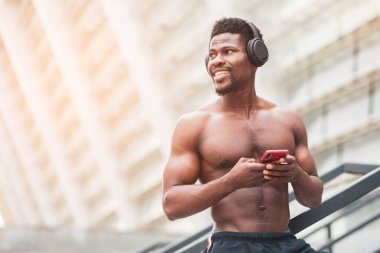 Afrikalı Amerikalı atletik adam yazın açık havada müzik dinliyor, tişörtsüz bir sporcu akıllı telefonu tutuyor ve fotokopi odasına bakıyor.
