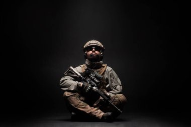 Amerikan askeri teçhizatlı ve tüfekli bir asker yerde oturur ve yere bakar, komando siyah bir arka planda düşünür ve üzülür.
