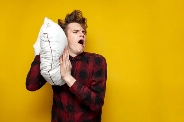 young sleepy tired guy yawns and holds a pillow on yellow isolated background, bad morning, insomnia concept
