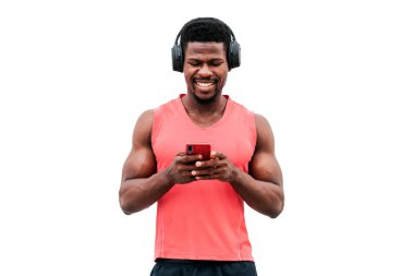 Afro-Amerikan atletik adam elinde bir akıllı telefon tutuyor ve beyaz izole bir arka planda müzik dinliyor, kulaklıklı bir adam telefona bakıyor ve gülümsüyor.