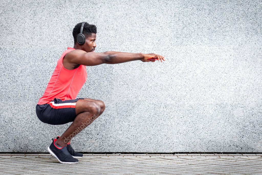 african american sports guy working out with headphones outdoors against wall, athletic man crouches and does physical exercises, copy space