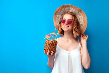 Güneş şapkalı ve gözlüklü bir kız mavi izole edilmiş bir arka planda tropikal ananas kokteyli içiyor. Egzotik içkisi olan bir kadın tatil köyünde dinleniyor, yaz konseptinde.