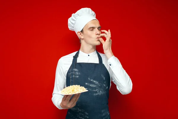 young chef holds a dish on a red isolated background and shows a sign of good taste, a kitchen worker in uniform cooked pasta