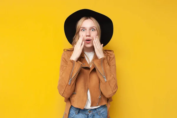Chockad flicka i en höst jacka och hatt är förvånad över en gul isolerad bakgrund — Stockfoto