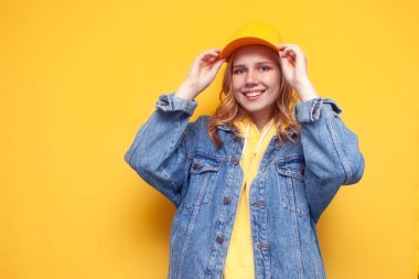 Kot ceketli genç kız sarı arka planda bir şapka takıyor ve gülümsüyor.