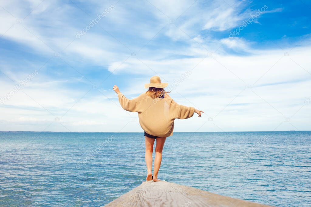 girl stands on a pier against the background of the sea and looks at the horizon, a woman in a sun hat waves her hands against the background of the sky, freedom concept