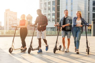 Irklar arası bir grup genç arkadaş şehirde elektrikli scooter kullanıyor ve iletişim kuruyor. Çok ırklı gençler elektrikli araçlar kullanıyor ve konuşuyorlar.
