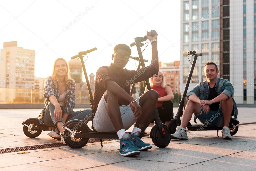 interracial group of young friends sitting with electric scooters in the city and smiling, multiracial youth use electric transport, eco transport rental concept