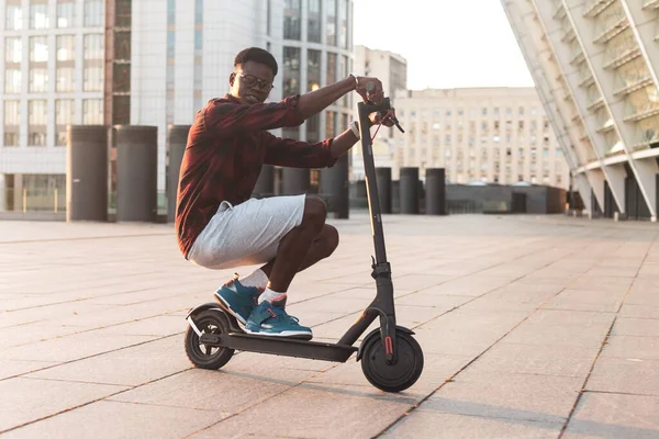 Komik afro-amerikalı adam şehirde elektrikli scooter kullanıyor ve gülümsüyor, öğrenci eko-taşıma kullanıyor, geleceğin ulaşımını kullanıyor.