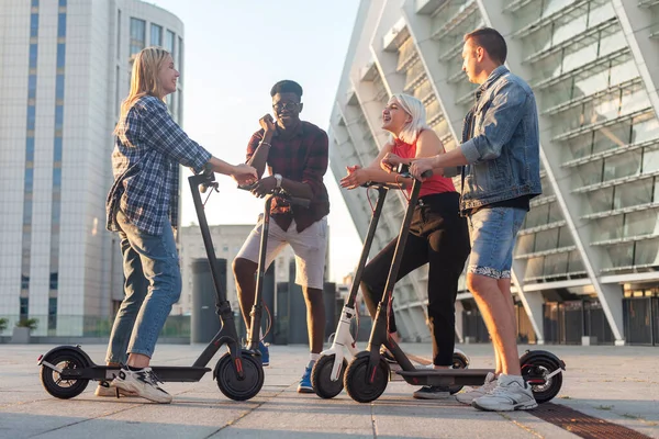 Irklar arası bir grup genç arkadaş şehirde elektrikli scooter kullanıyor ve iletişim kuruyor. Çok ırklı gençler elektrikli araçlar kullanıyor ve konuşuyorlar.