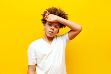 Beyaz tişörtlü yorgun Afro-Amerikan kıvırcık çocuk üzgün ve sarı izole edilmiş arka planda sıkılmış, mutsuz ergen elleriyle alnını siliyor ve sorunlarından dolayı iç çekiyor.