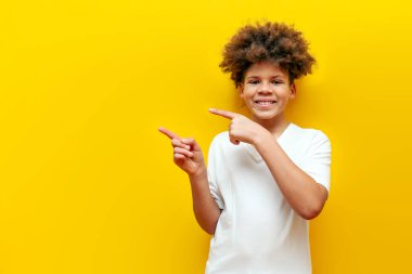 Neşeli kıvırcık Afrikalı Amerikalı çocuk sarı izole arka planda fotokopi alanını işaret ediyor. Beyaz tişörtlü ve boş alan reklamı yapıyor.