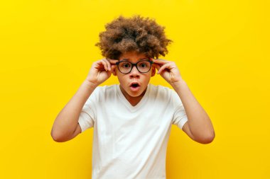 Şaşırmış kıvırcık saçlı gözlüklü Amerikalı çocuk sarı izole edilmiş arka planda kameraya bakıyor beyaz tişörtlü genç şaşkınlıkla fotokopi odasına bakıyor.