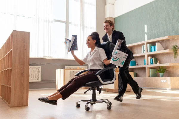 Ofis çalışanları iş kıyafetleri içinde eğleniyor ve ellerini kaldırarak sandalyelere biniyor. Afrikalı Amerikalı kadın Avrupalı genç adamla başarıyı kutluyor ve işindeki zaferle seviniyor.
