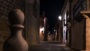 A narrow alleyway of a medieval town by night. The alleyway is dark and quiet, with only a few lights illuminating the area