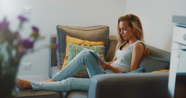 Imagens de alta qualidade 12bit de menina bonita usando Tablet PC sentado no sofá em casa. Mulher navegando na web, redes sociais, procurando algo na loja online . — Vídeo de Stock