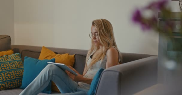 Hochwertige 4k 12bit Filmmaterial von schönen Mädchen mit Tablet-PC sitzt auf der Couch zu Hause. Frau surft im Internet, soziale Netzwerke, sucht etwas im Online-Shop. Filmischer Look — Stockvideo