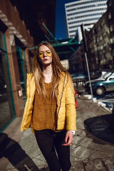A young European woman, traveling, with long blond hair, wearing a yellow jacket, yellow sunglasses walking down the city center street, street shooting. Even light.
