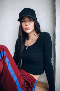 Bir kap ve spor giyim esmer kız portresi. Doğal ışık. Kırmızı eşofman altı, siyah en iyi, siyah başlığı.